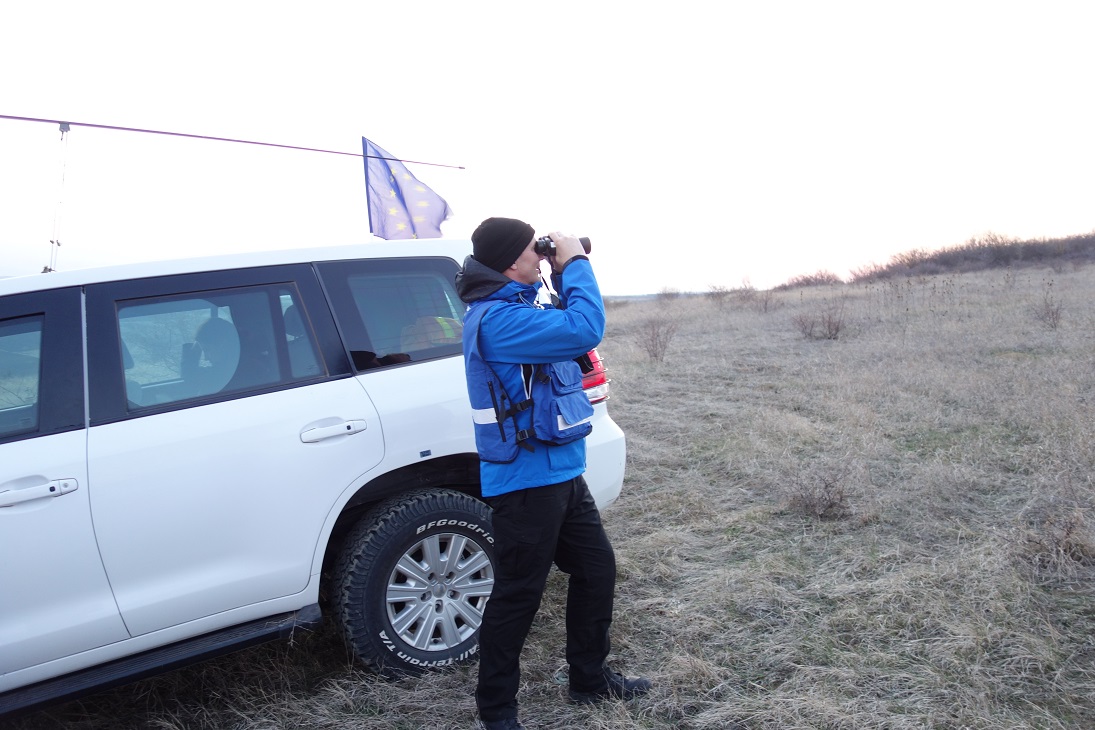 Mies sinisessä takissa ja mustissa housuissa seisoo valkoisen auton edessä ja kiikaroi kaukaisuuteen